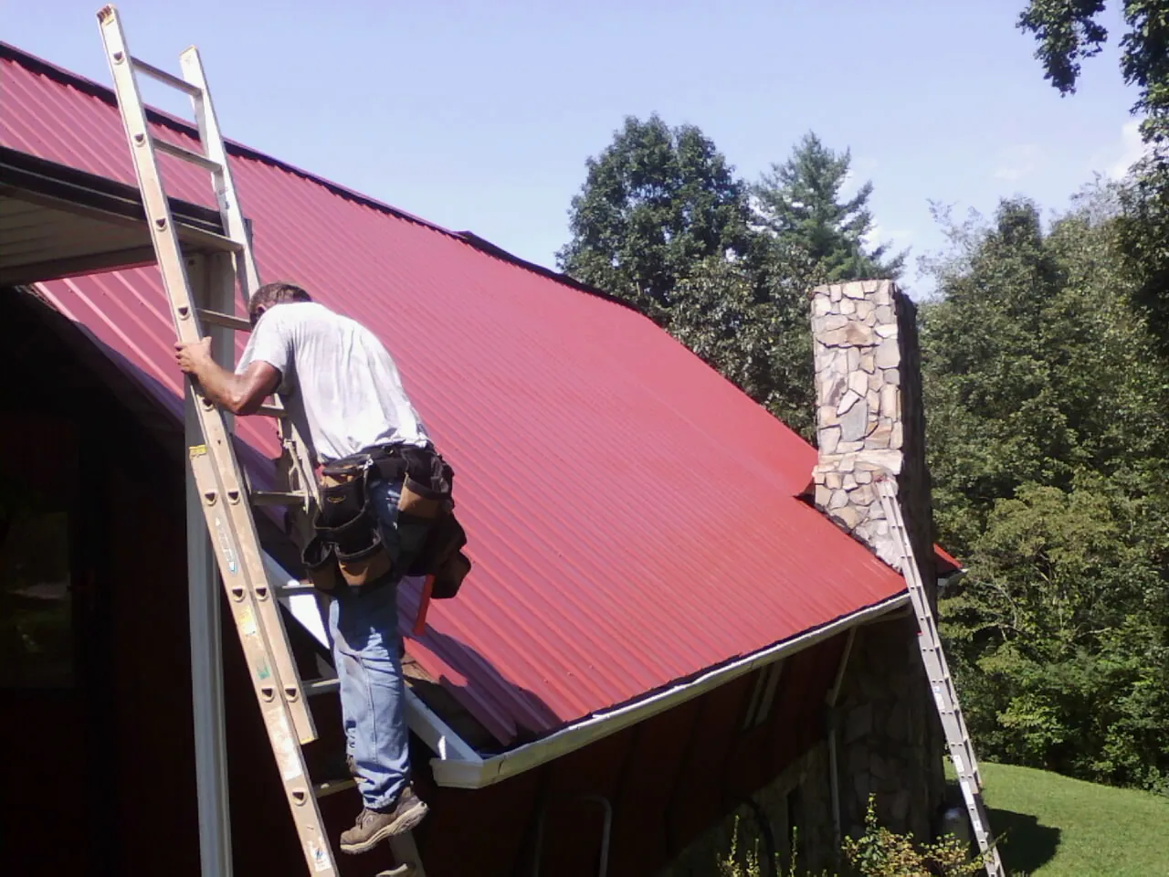 Roofer on a ladder beside a red metal roof and stone chimney — Soffit & Fascia Repair by Roofing Brothers in Evant